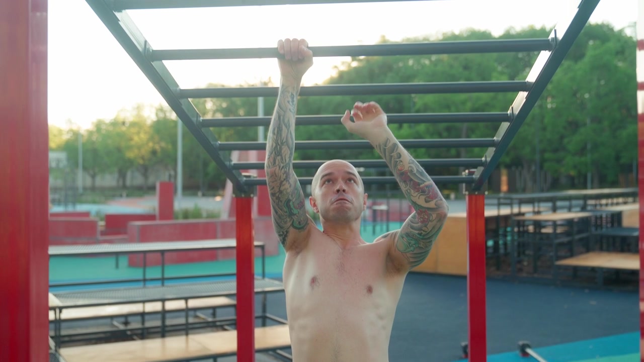 Uomo tatuato a torso nudo che si esercita alle sbarre orizzontali in una palestra all'aperto.