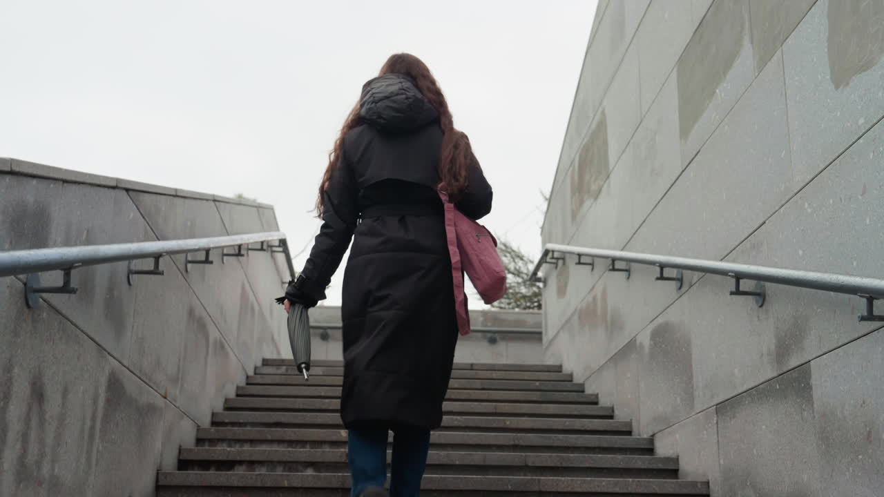mujer blanca subiendo escaleras de hormigón, cielo cubierto, abrigo negro grueso, bolso rosa colgado al hombro, pasamanos de metal a ambos lados, paso subterráneo de hormigón, pasos lentos y constantes, viaje solitario, reflectante