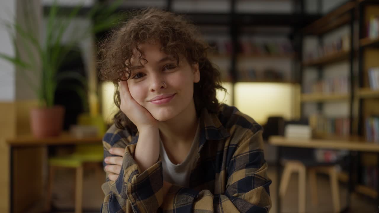 Portrait of a happy student girl with curly hair posing while sitting at a table in the library