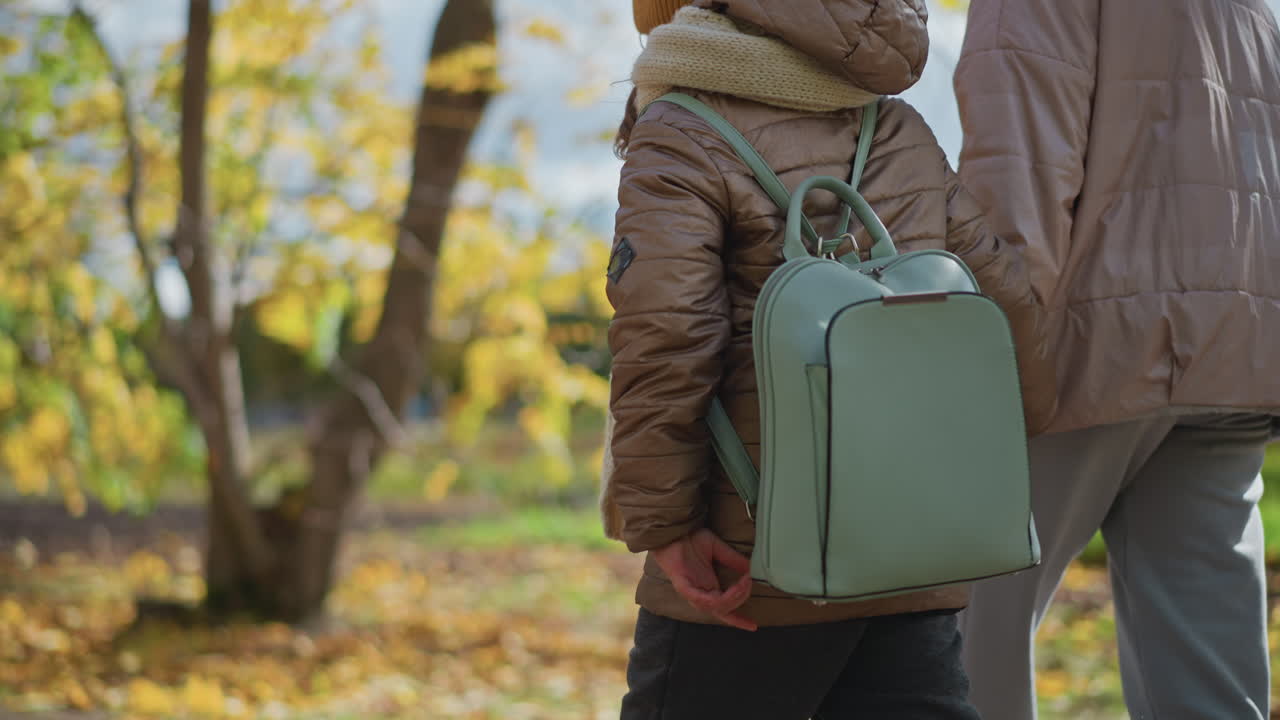 low angle side view of mother walking with child while conversing along dirt path strewn with colourful autumn leaves in open field near tree trunk wearing jackets joggers and sneakers