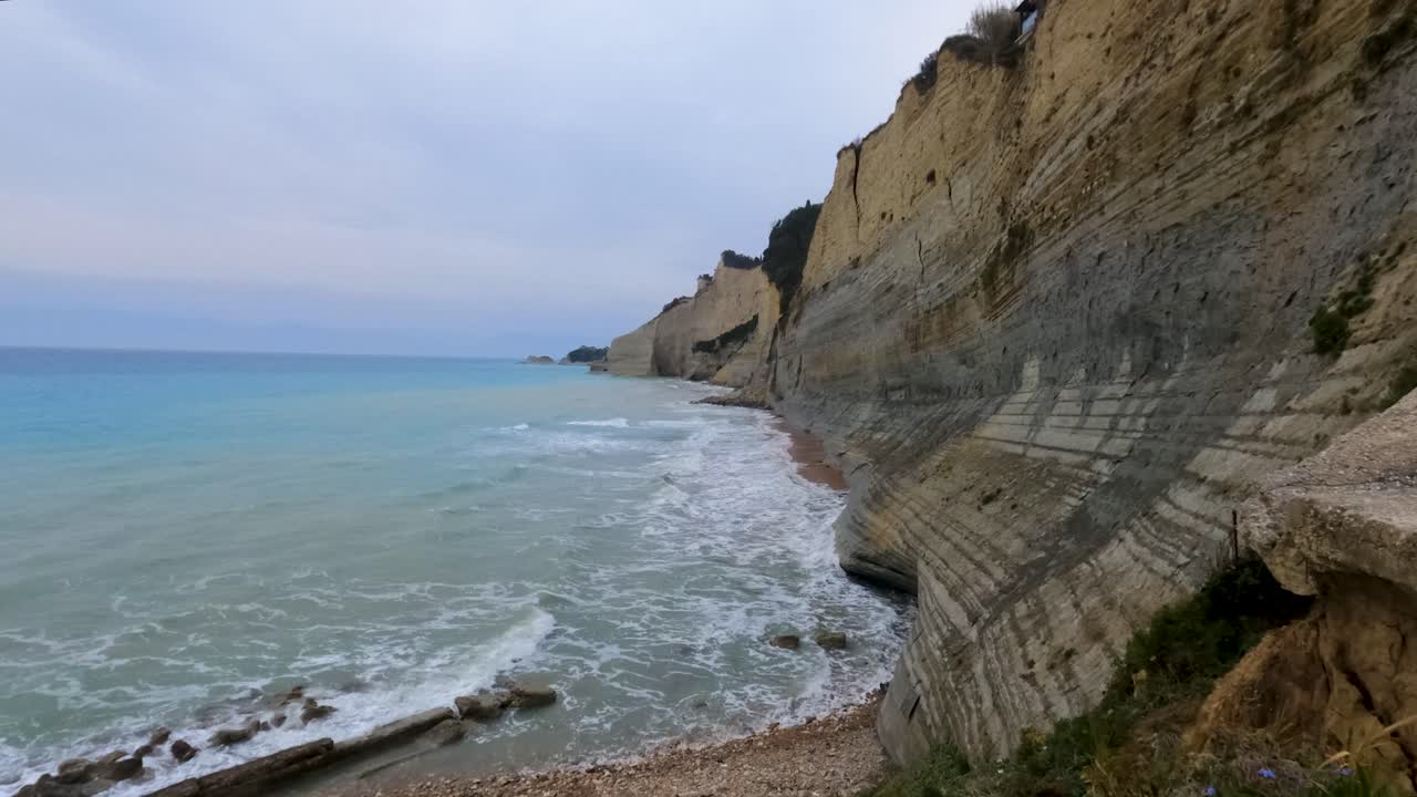 Ripide scogliere di argilla a picco sul mare turchese, spiaggia di Loggas a Corfù, formazione geologica della costa, onde che si infrangono sulla riva ghiaiosa, strati rocciosi erosi, maestoso paesaggio naturale