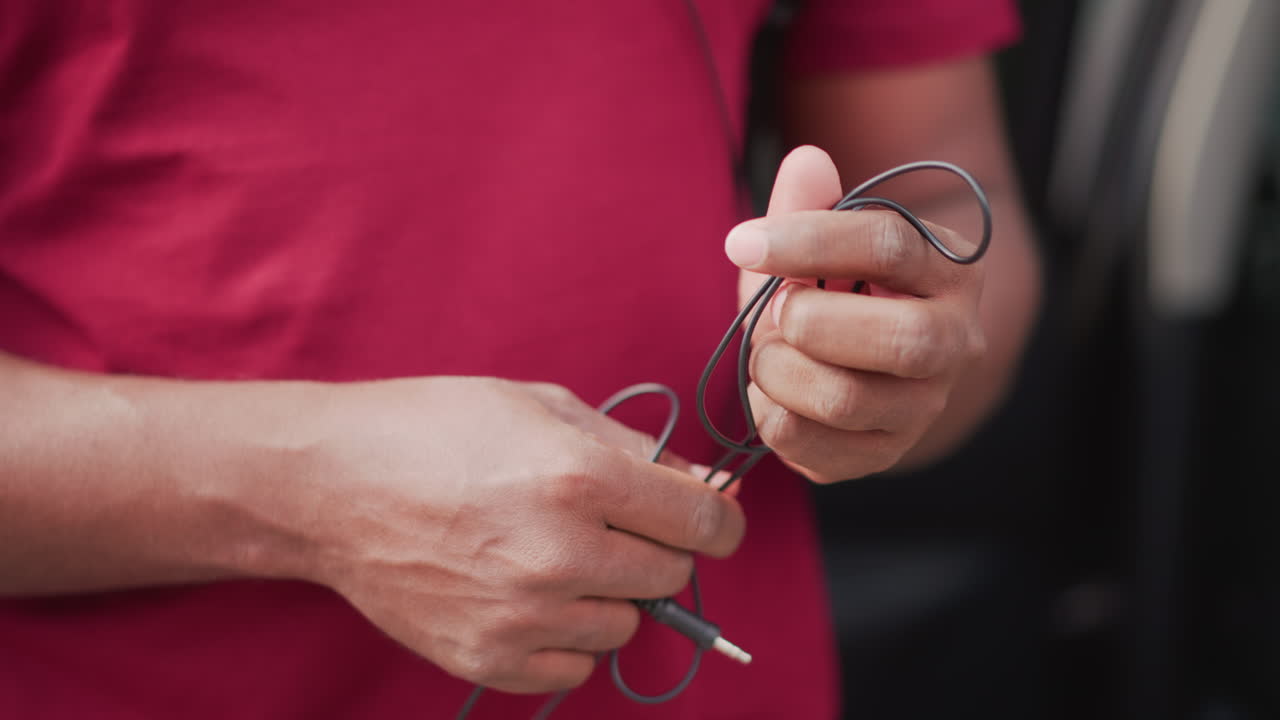 Tecnico del suono ripara il cavo aggrovigliato delle cuffie, primo piano illuminato dal sole di mani esperte in maglietta rossa che testano un connettore vicino a un veicolo, soundcheck metodico e allestimento dell'attrezzatura, dita precise.