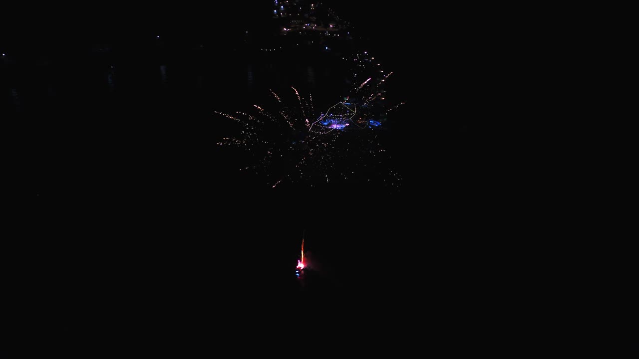 Una rotazione aerea con punto di interesse, attorno a fuochi d'artificio che esplodono di notte dal mare, vicino a un molo