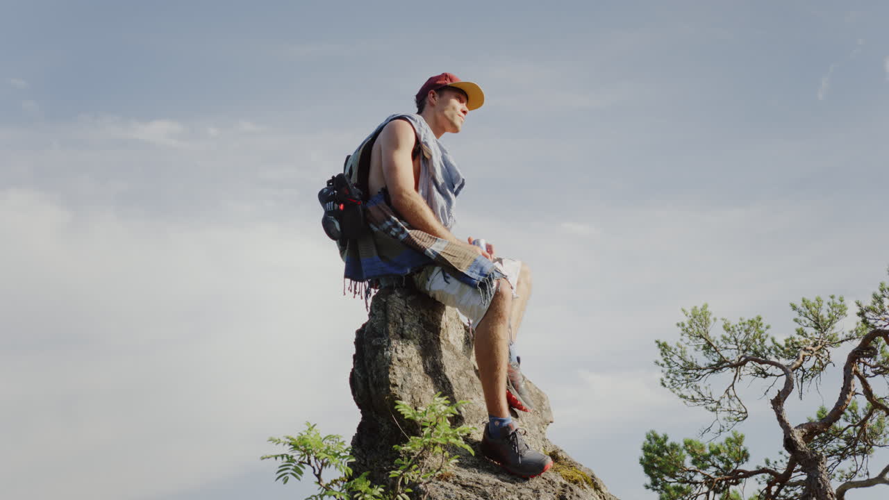 handsome guy looking around sitting on a mountain rock, blue sky air in background, tree branch in the cinematic shot