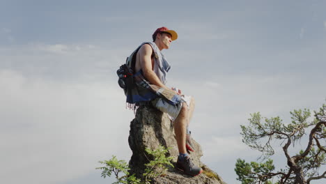 Un-Tipo-Guapo-Mirando-A-Su-Alrededor-Sentado-En-Una-Roca-De-Montaña,-Aire-De-Cielo-Azul-En-El-Fondo,-Rama-De-árbol-En-La-Toma-Cinematográfica