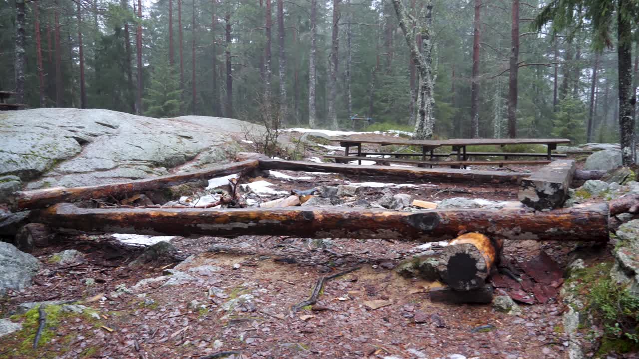 Picnic tables and logs lashed together to form sitting benches around a fire pit at a clearing in the forest on a damp and misty day