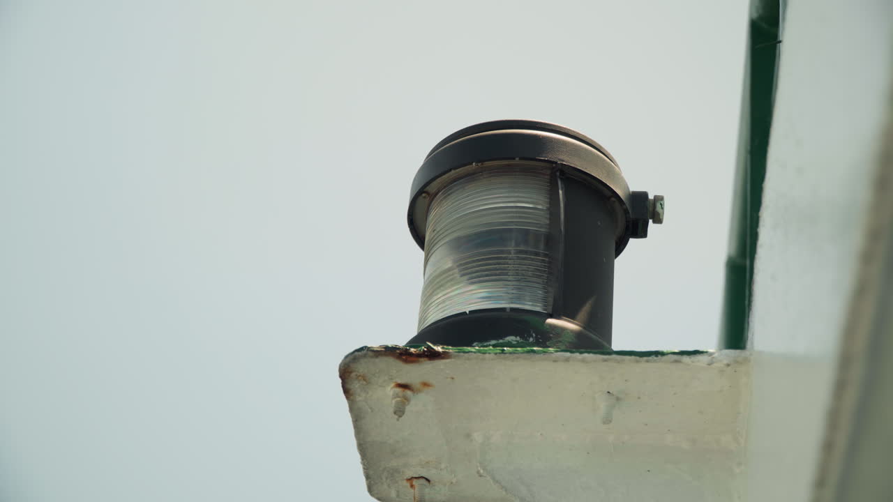 Luce arrugginita sul tetto contro un cielo pallido, primo piano di un apparecchio metallico invecchiato montato su un cornicione di cemento con alloggiamento in vetro e sigillante verde, sfondo nuvoloso e smorzato che evoca un'atmosfera industriale, corrosione