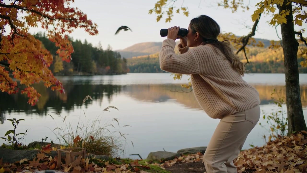 Watching woman in tan sweater rising at lakeside blanket after bird flies, tracking with binoculars