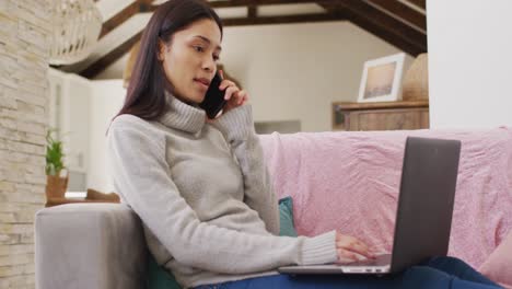 Biracial-woman-talking-on-smartphone-and-using-laptop-in-living-room