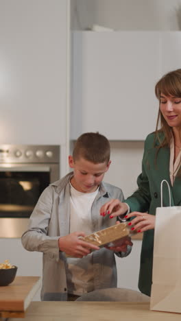 smiling pleasant woman in business suit takes out food from bags. schoolboy helps mom taking waffles, cookies and cupcakes. preparing for family dinner