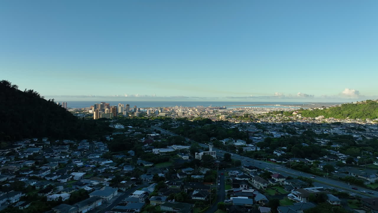 Suburb neighborhood of Honolulu and downtown Skyscraper in distance. Sunset time in America against blue sky and pacific ocean. Aerial rising wide shot. Traffic on highway.