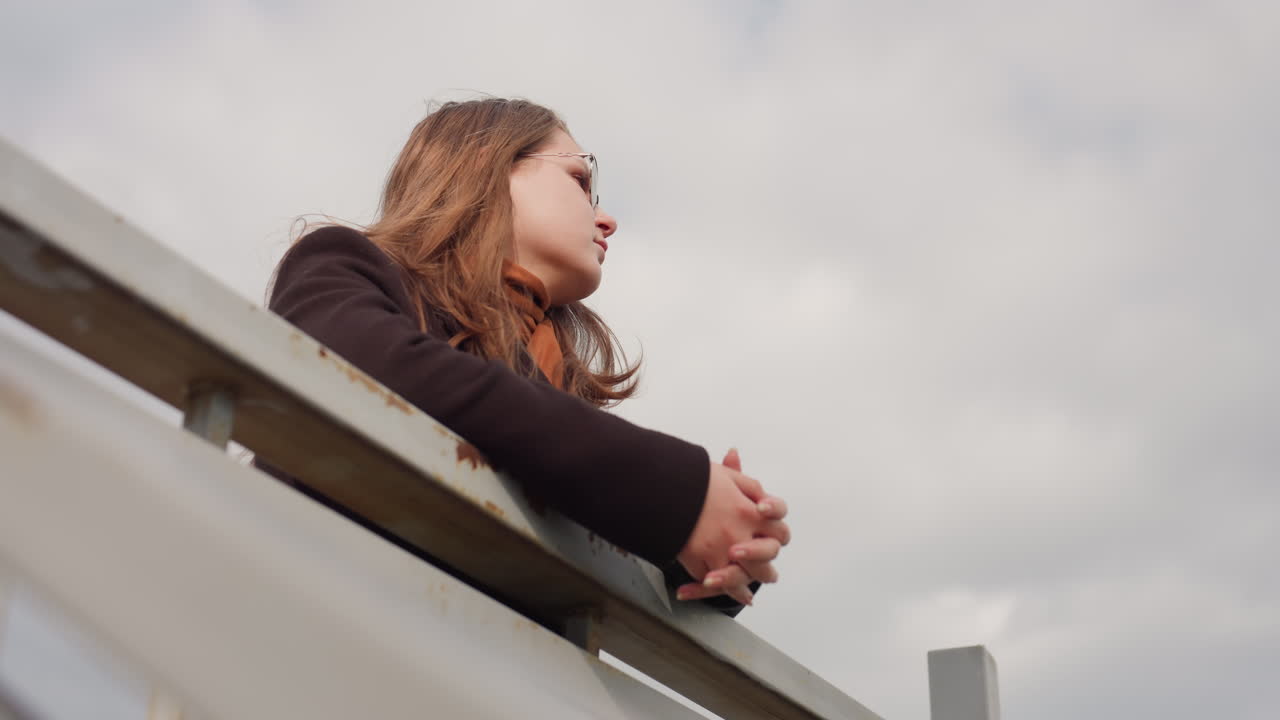 Elegant woman wearing wool coat and eyewear stands outdoors near security railing, enjoying fresh air under dusty sky expressing calmness, elegance, and peaceful reflection in natural daylight scene