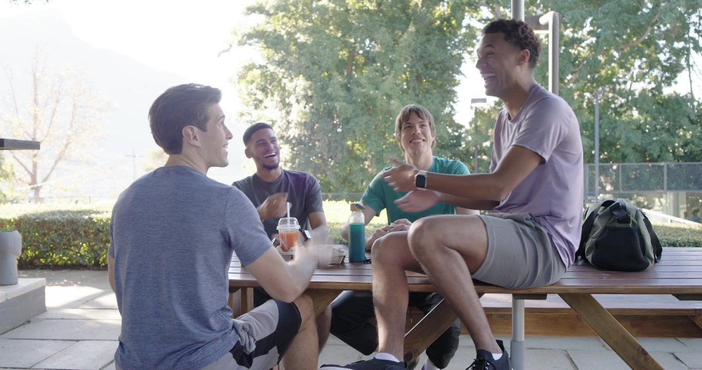 Diverse mannelijke studenten eten aan campustafel nadat een student op een bankje gebaart en een blauwe fles doorgeeft.