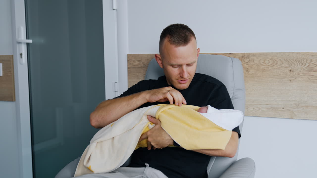 Happy father sits in armchair holding his newborn baby. New-made parent taking care of a child after birth in hospital.