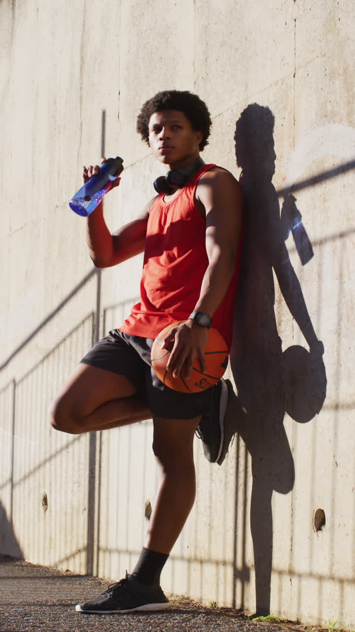 disparo vertical de un hombre biracial apoyado en la pared, bebiendo agua, sosteniendo una pelota de baloncesto