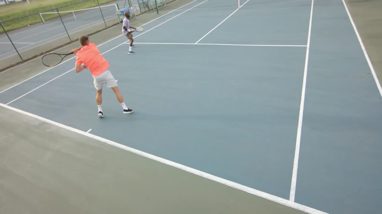 Dos amigos diversos jugando dobles y devolviendo la pelota en una cancha de tenis al aire libre, a cámara lenta.