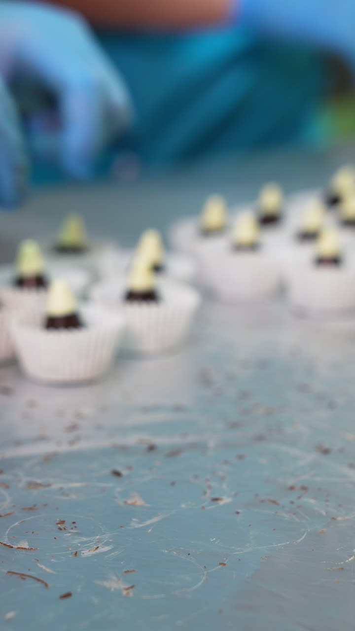 Confectioner takes little chocolate cones with white tops and puts them in paper. Packaging ready-made sweets at factory. Blurred backdrop. Vertical video