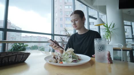 young woman eating a fresh salad at a wooden table in a modern restaurant with large windows, natural light, and urban views. relaxed dining scene