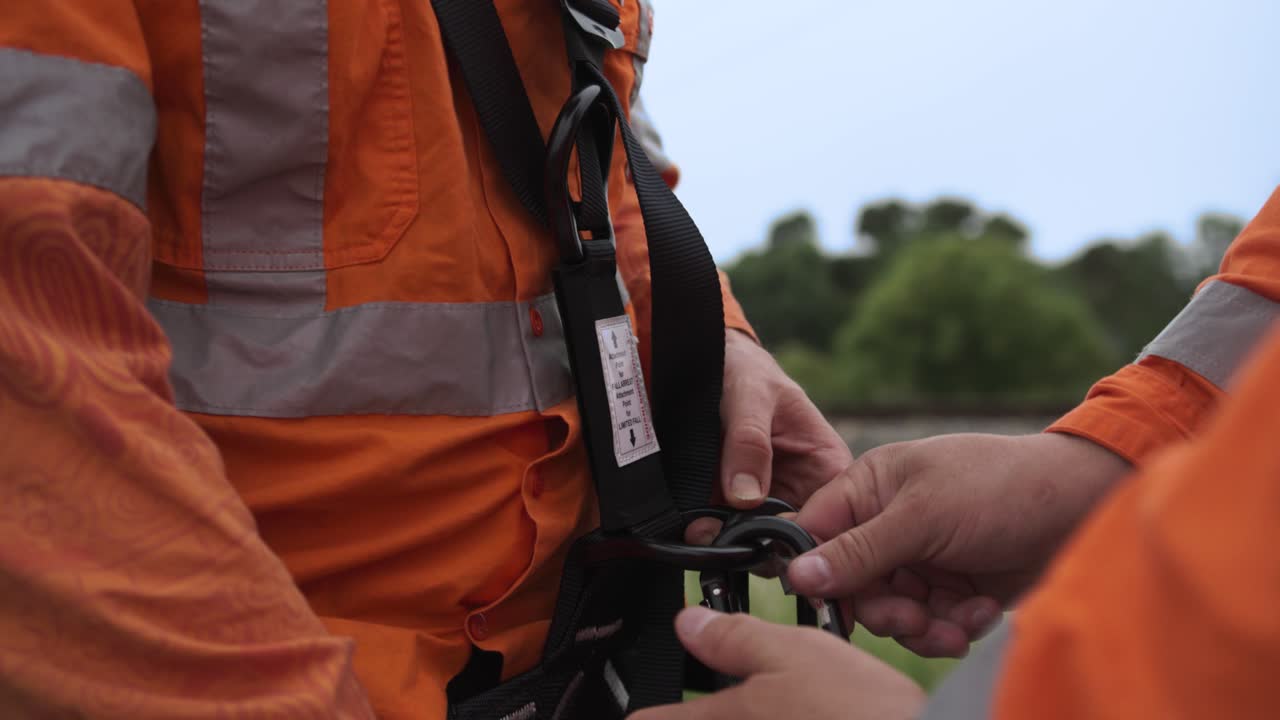 Close-up of a railway worker in high-visibility orange clothing fastening a safety harness with carabiners near a railway corridor