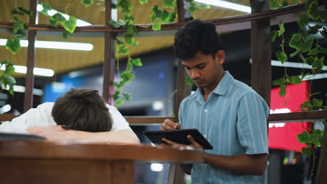 blurry view of tall stone pillars and hanging flower garlands framing light skinned boy with hoop earring gazing beyond glass partition under soft golden glow in serene indoor garden setting