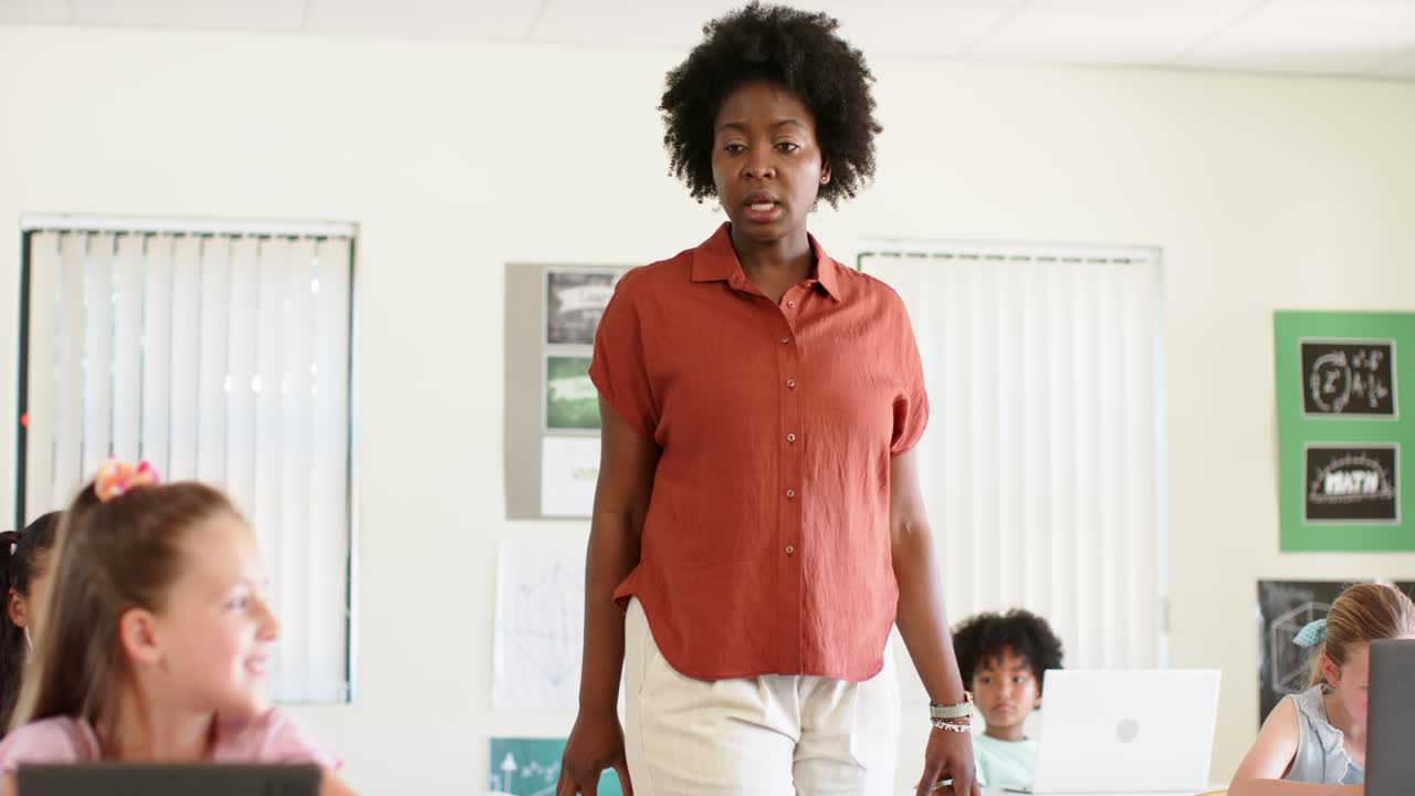 African American female teacher walking down center aisle in classroom checking tablets and laptop