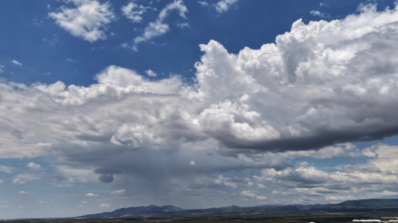 4K drone timelapse capturing fast-moving cumulonimbus rainclouds and precipitation shafts. Heavy atmospheric weather surge over a vast mountain and mesa canyon landscape against a deep blue sky.