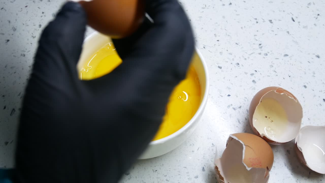 A woman wearing a black rubber glove was breaking some raw eggs into a white bowl. This process is carried out carefully to maintain the shape of the yolk.