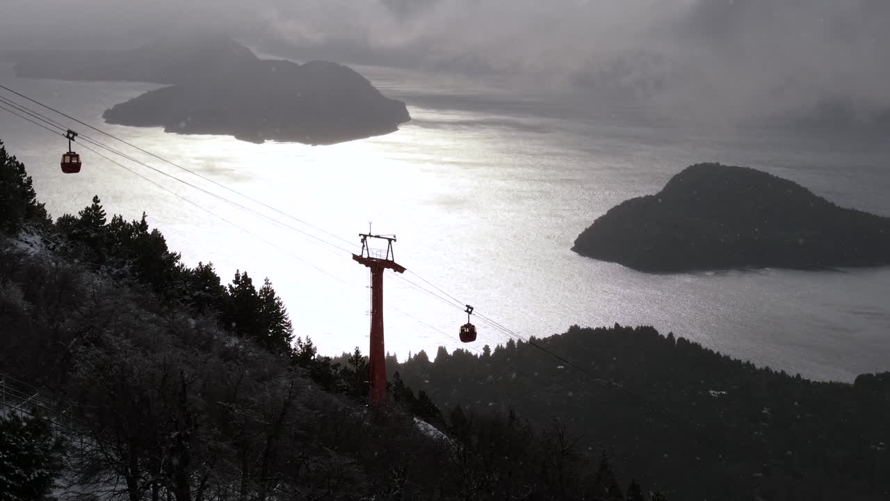 Snowy Cerro Otto cable car tower above shimmering lake and forested islands under dramatic winter sky