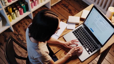 woman working from home on laptop in a cozy candle-making business workspace.