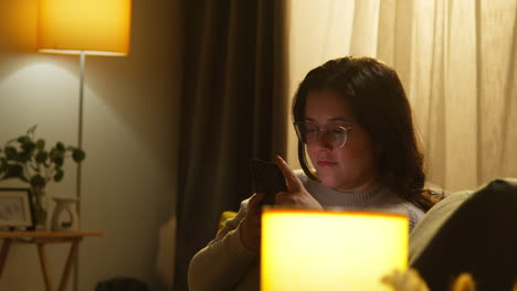 mujer joven en casa sentada en el sofá con el teléfono móvil desplazándose por internet o redes sociales en la noche 5