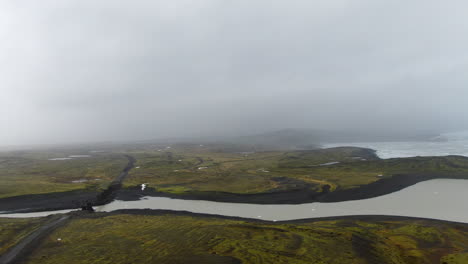 Luftaufnahme-Der-Isländischen-Landschaft-Mit-See-Und-Grünem-Gras-Und-Weiß-grauem,-Wolkigem,-Nebligem-Himmel-Und-Schotterstraße