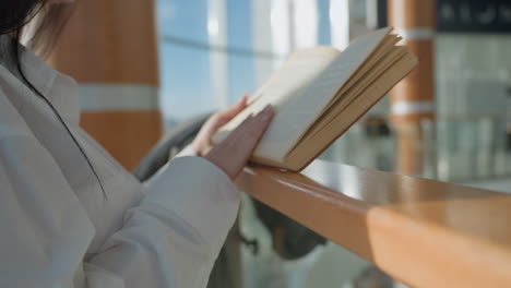 close up of young woman in white shirt reading open book while leaning on wooden rail in brightly lit indoor space, hands gently holding pages as sunlight highlights scene with soft shadows