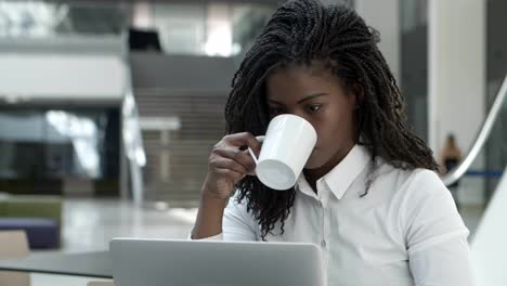 Focused-young-woman-drinking-coffee-and-looking-at-laptop