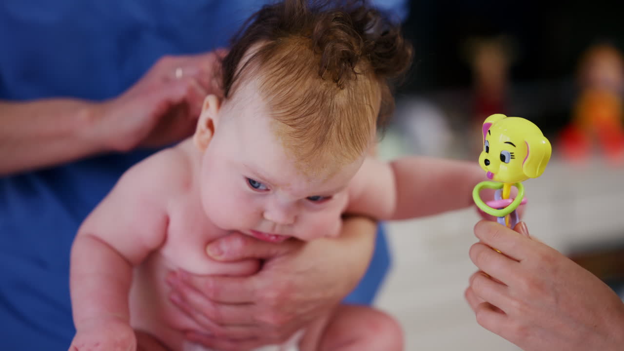 Woman trying to distract a crying baby lying on a blanket during massage with a toy