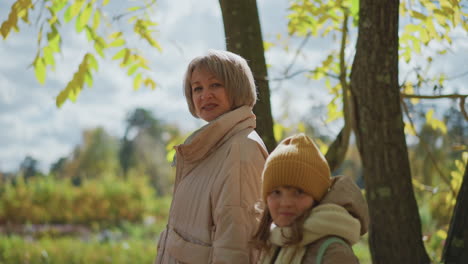 portrait of mother and daughter smiling warmly at camera during autumn walk under sunlit trees with leaf litter on ground soft focus background