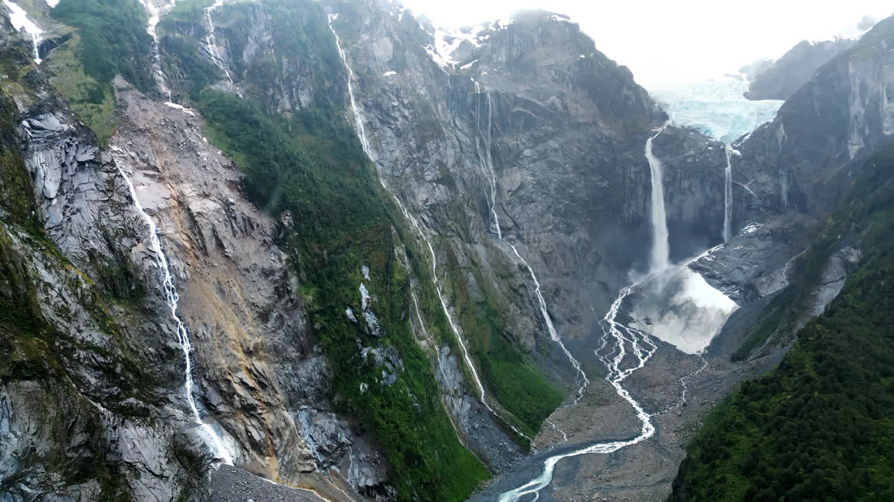 Aerial drone view of the stunning Ventisquero Colgante glacier waterfall cascading down steep cliffs surrounded by lush green forest in slow motion