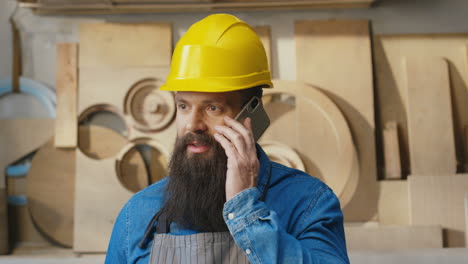 portrait of caucasian bearded man in apron and helmet talking on the phone in carpentry workshop