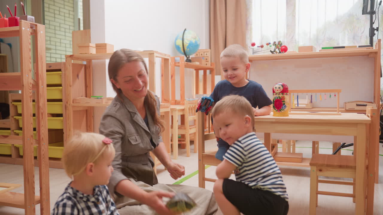 Aufsichtsperson mit Kleinkindern im Klassenzimmer, die auf dem Boden sitzen und einer Schildkröte zusehen, die sich bewegt. Ein Junge mit einem gestreiften Oberteil beugt sich interessiert näher, ein Mädchen hält ein Stofftier, ein anderes Kind sitzt in der Nähe und hält ein Spielzeugauto.