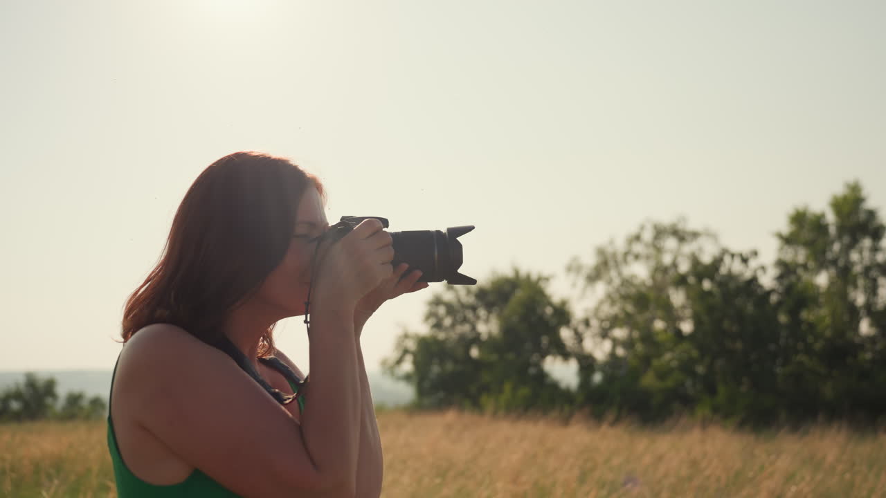 woman in green dress walks through golden field holding camera as sunlight glows behind her while gazing toward open landscape and distant river evoking freedom exploration and quiet reflection