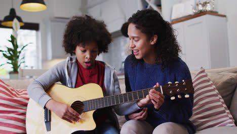 Mixed-race-mother-and-daughter-sitting-on-couch-playing-guitar