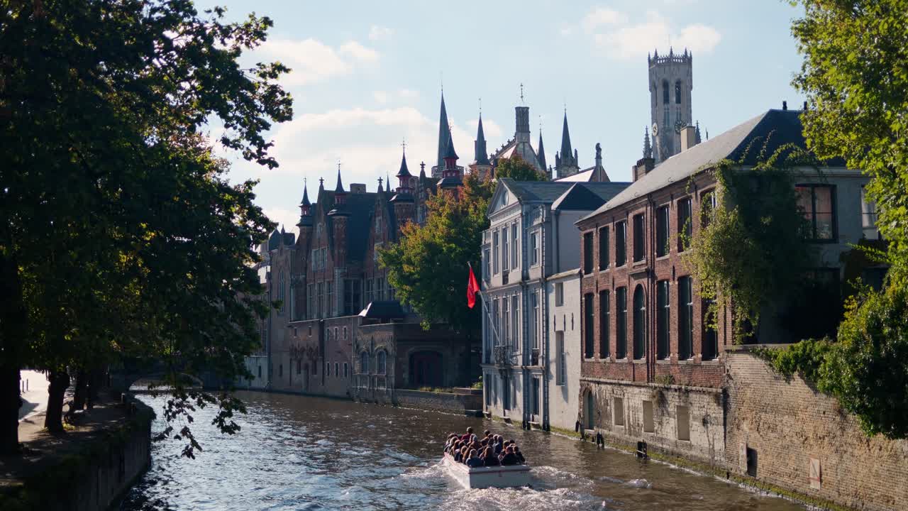 A boat with tourists cruises along a canal in scenic Bruges with historic buildings and trees