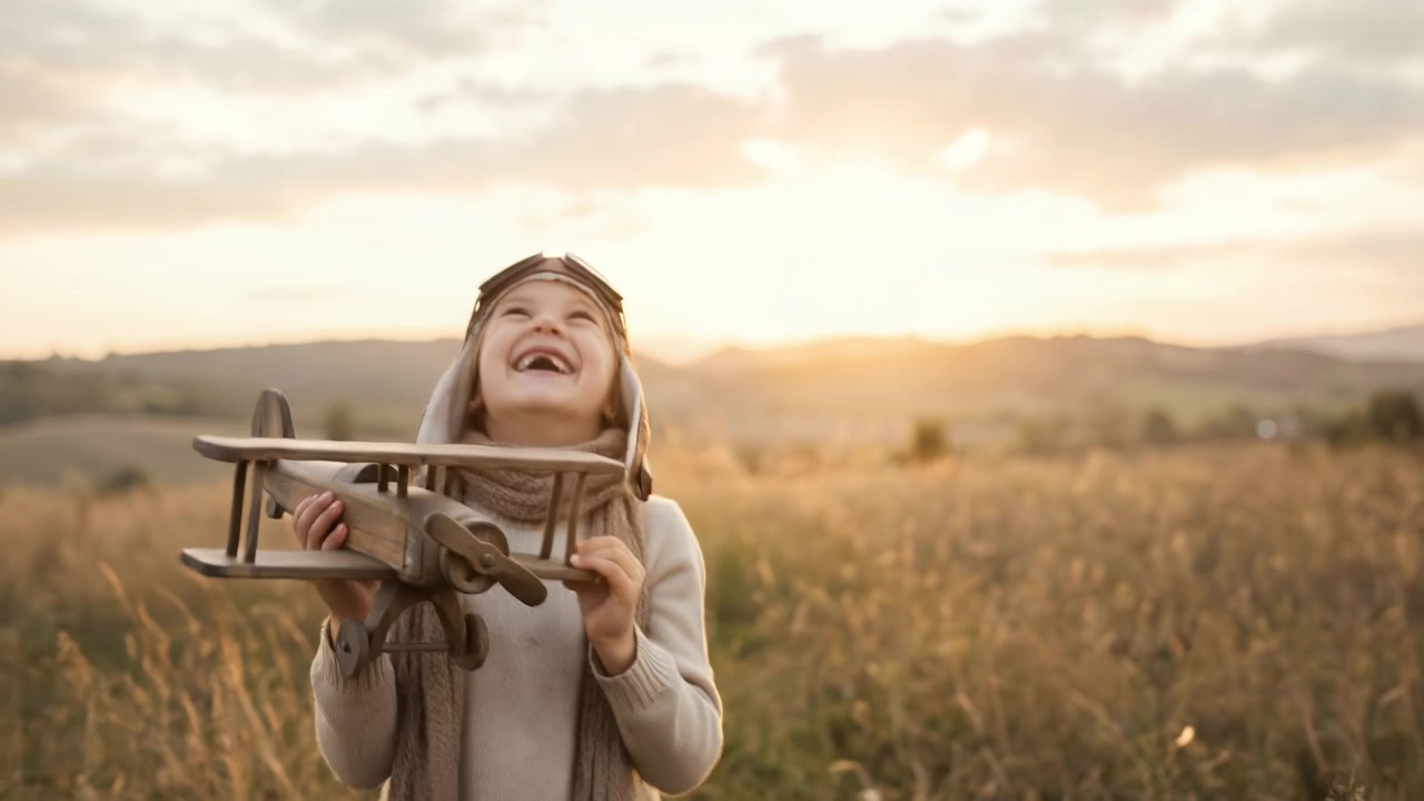 Menino jovem com equipamento de aviador olhando atentamente para um avião de brinquedo de madeira em um campo ensolarado