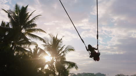 travel woman swinging over jungle at sunrise enjoying exotic vacation sitting on swing with sun flare shining through palm trees in tropical rainforest holiday lifestyle freedom