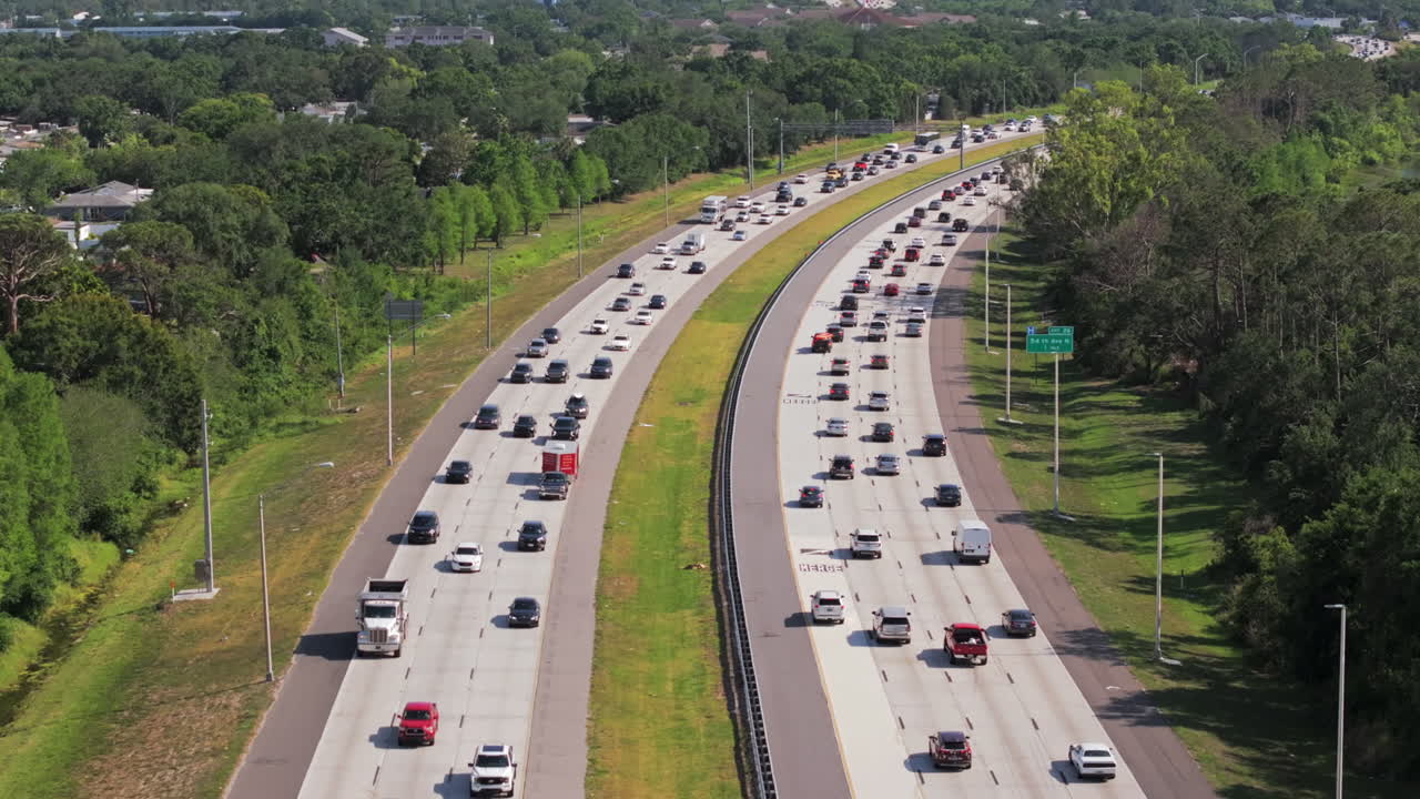 Aerial footage captures vehicle traffic driving a divided highway cutting through green trees beyond a suburban neighborhood showcasing modern highway infrastructure.