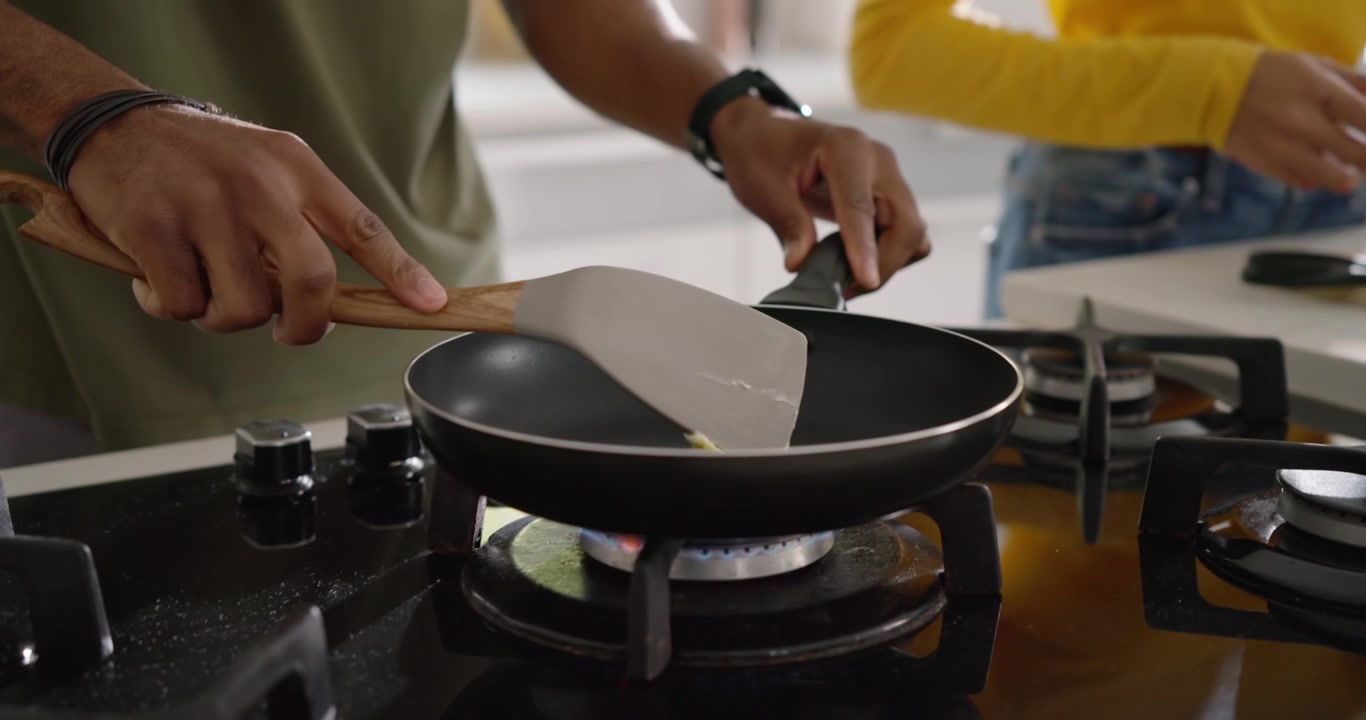 Diverse couple peeling carrots with peeler on board as man entering, heating pan for cooking