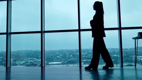 woman in loose suit walks along the panoramic windows in the office. silhouette of female employee. low angle view.
