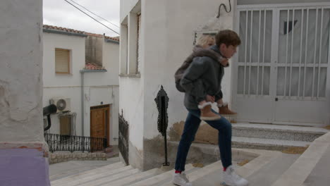 young boy carrying a little girl on his back while walking up stairs in a village street