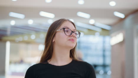 primer plano de una joven elegante con gafas y camiseta negra disfrutando de un paseo relajado en un centro comercial moderno, con transeúntes borrosos y luces bokeh brillantes en el fondo