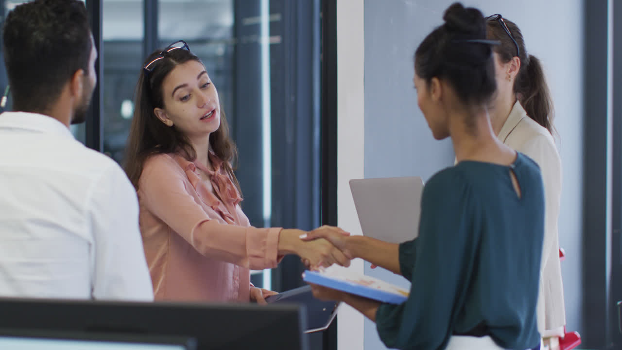 dos mujeres de negocios diversas de pie estrechando la mano durante una reunión con colegas