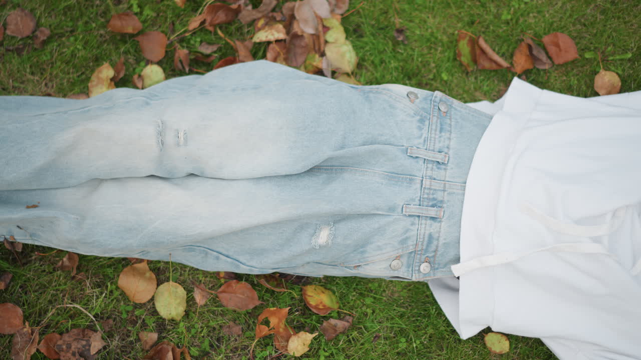 White girl feet in slippers outdoors, textured denim trousers, fallen leaves on grass, cozy socks and slip slippers, fashion blogger relaxing on lawn, intimate lifestyle closeup, warm autumn light